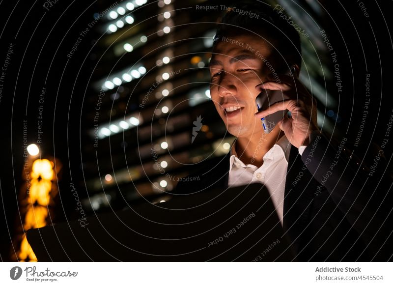Asiatischer Geschäftsmann am Laptop und im Gespräch mit dem Smartphone auf einer dunklen Straße Mann Telefonanruf diskutieren Gebäude Lächeln Nacht Glück