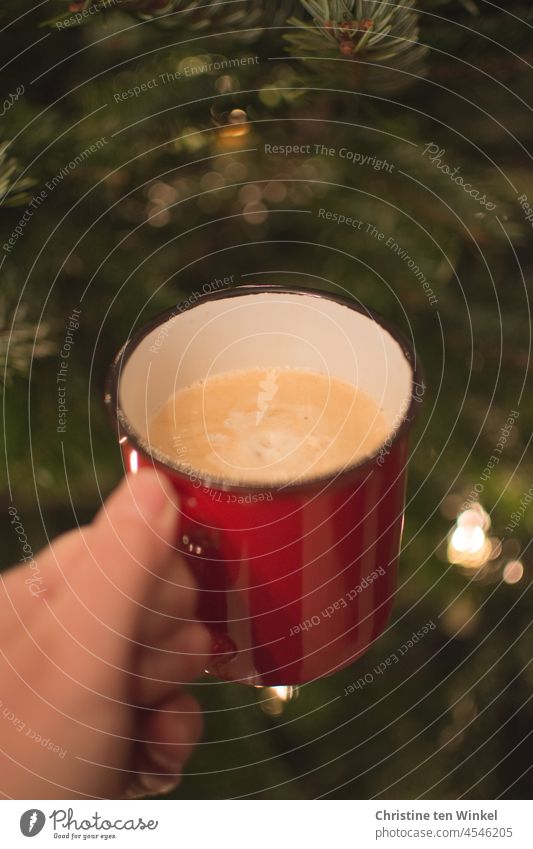 Eine Hand hält einen roten Becher mit Cappuccino. Im Hintergrund Tannengrün mit glänzenden Lichtern. Adventskaffee. Kaffeebecher festhalten Tasse Weihnachtsbaum