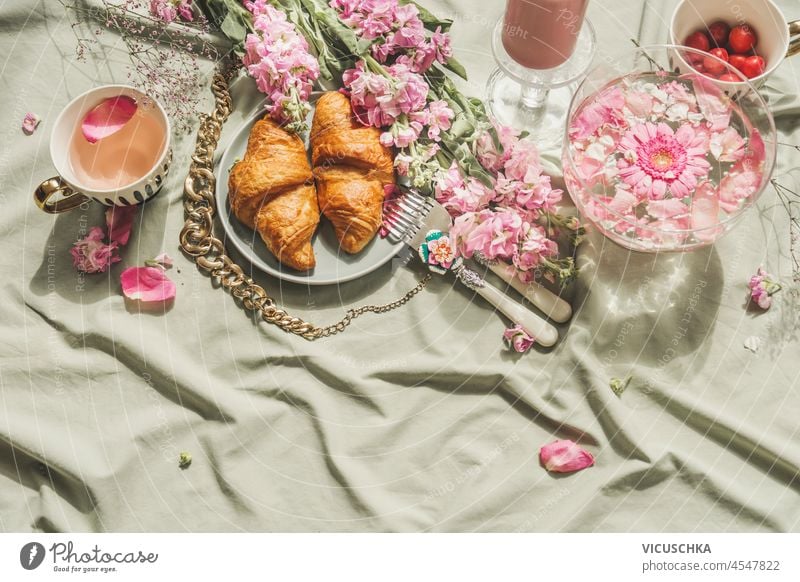Sommerpicknick mit französischem Croissant, Kirschblütentee und frischen Kirschen auf hellem Textilhintergrund. Romantische Frühstücksszene mit rosa Blütenblättern, Speisen und Getränken. Ansicht von oben.