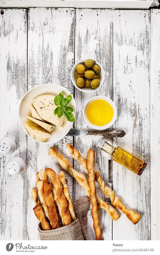 Knusprige Grissini-Brotstangen. Traditionelles italienisches Weizenbrot mit Knoblauch, Käse und Sesam. Lebensmittel-Stillleben auf einem weißen, rustikalen Hintergrund. Olivenöl, Ziegenkäse und grüne Oliven. Ansicht von oben