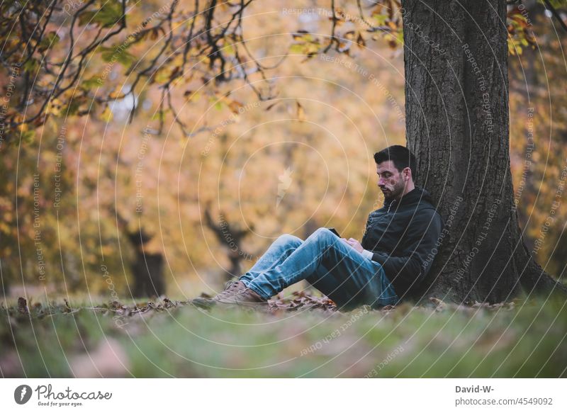 Mann sitzt draußen in der Natur an einem Baum und genießt die Ruhe ausruhen genießen Handy entspannen Auszeit Herbst herbstlich Abstand