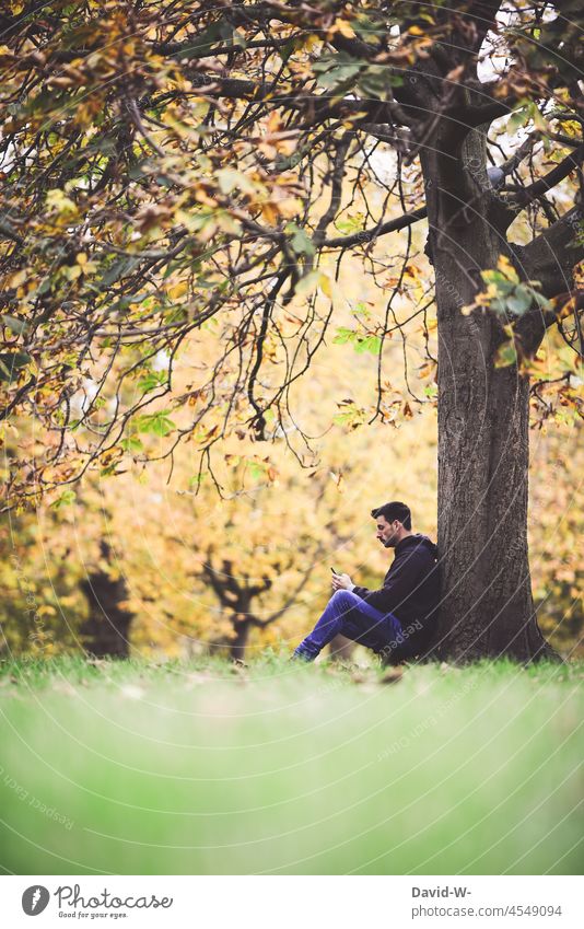 Mann in der Natur schaut auf sein Handy draußen benutzen Smartphone Erreichbarkeit Mobile Lifestyle Internet Mitteilung vertieft lesen Tunnelblick Herbst Baum