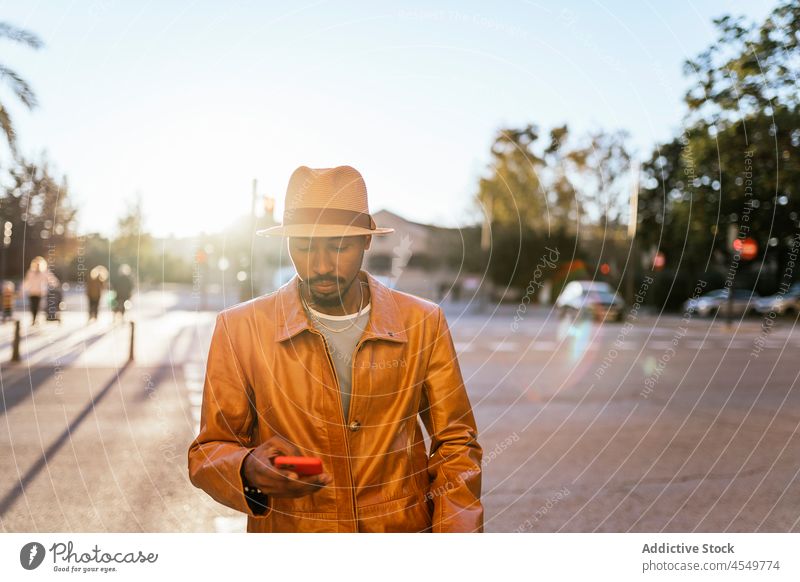 Stilvoller schwarzer Mann, der auf der Straße sein Smartphone durchsucht Browsen online Mode trendy Textnachricht Anschluss Afroamerikaner Hut Surfen benutzend