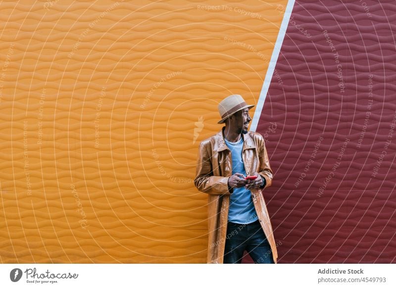 Fröhlicher schwarzer Mann, der in der Nähe der Wand sein Smartphone durchsucht Browsen online Stil Straße trendy Textnachricht Anschluss Afroamerikaner Hut