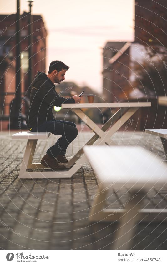 Mann sitzt draußen an einem Tisch eines Kaffees und benutzt sein Handy sitzen konzentriert Kaffeetrinken Kaffeepause Gastronomie Bank Kaffeetisch öffentlichkeit