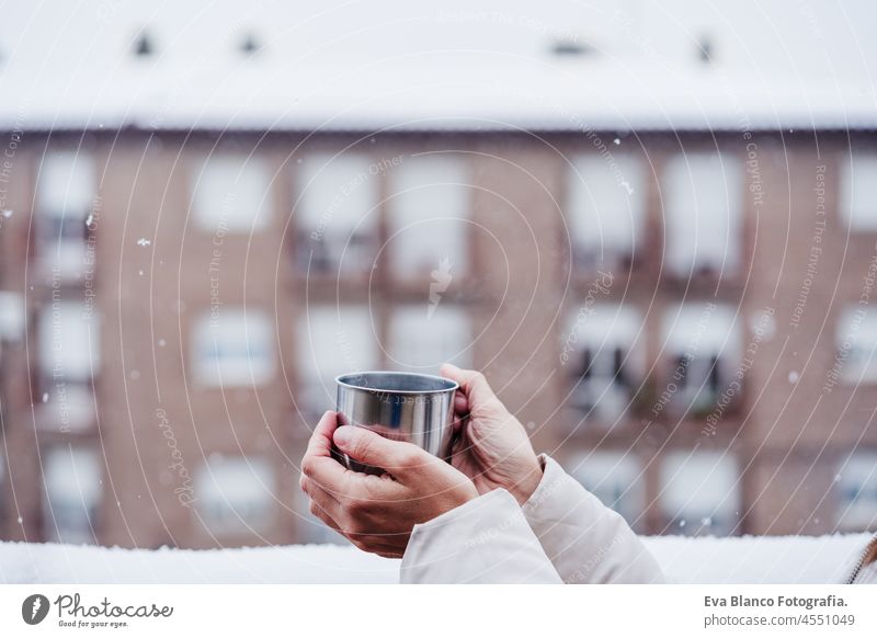 Seitenansicht der glücklichen Frau auf der Terrasse zu Hause, die eine Tasse heißen Kaffee genießt, Nachbarschaft Hintergrund. Schnee in der Stadt Winter