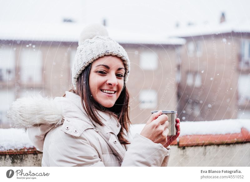 glückliche Frau auf der Terrasse zu Hause, die eine Tasse heißen Kaffee genießt, Nachbarschaft Hintergrund. Schnee in der Stadt Winter heimwärts Rauch