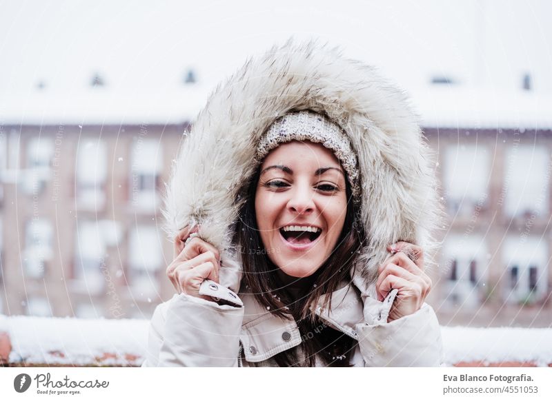 glückliche Frau auf Terrasse zu Hause genießen Schnee hält Kapuze mit Händen, Nachbarschaft Hintergrund. Schnee in der Stadt Kaukasier Großstadt Glück urban