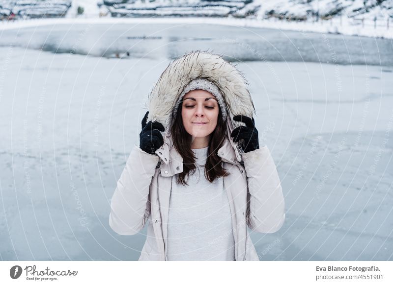 Porträt einer entspannten schönen kaukasischen Frau mit geschlossenen Augen, die einen Mantel mit Kapuze hält und vor einem zugefrorenen See in der Stadt steht. Lifestyle im Winter in der Stadt