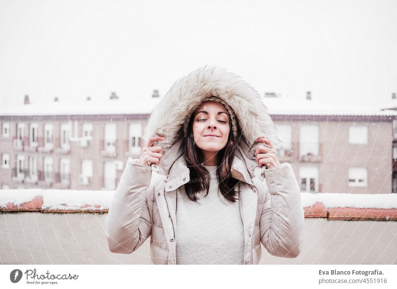 entspannte Frau mit geschlossenen Augen auf der Terrasse zu Hause genießt Schnee hält Kapuze mit den Händen, Nachbarschaft Hintergrund. Schnee in der Stadt