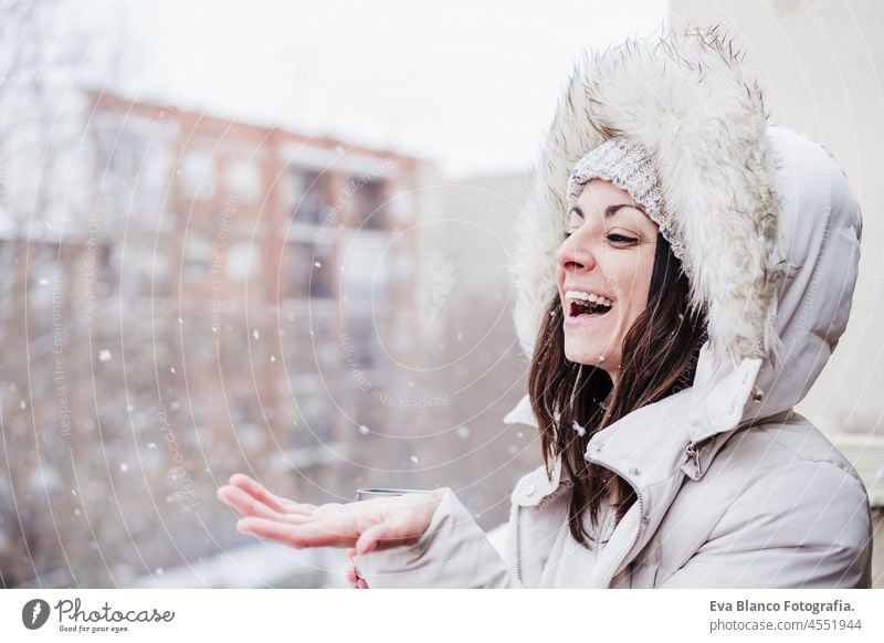glückliche Frau auf Terrasse zu Hause genießen Schnee hält Kapuze mit Händen, Nachbarschaft Hintergrund. Schnee in der Stadt Winter heimwärts Glück Kaukasier