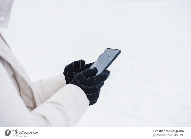 unerkennbare Frau, die im Winter in der Stadt spazieren geht, während es schneit, und dabei ihr Handy benutzt.winter lifestyle unkenntlich Schnee