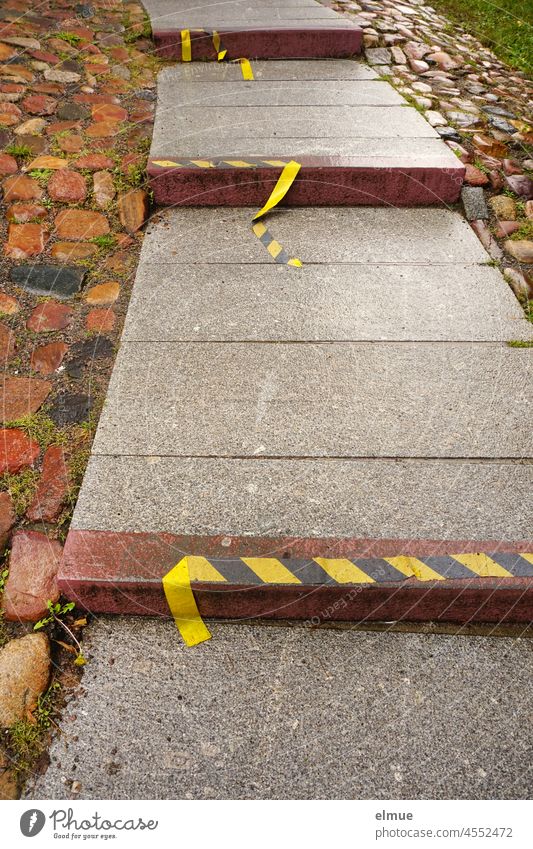 ein mit Natursteinen gesäumter Steinweg aus großen Platten und eingebauten Stufen, von denen sich das gelb-schwarze Markierungsband ablöst / Aufstieg / Bodenmarkierung