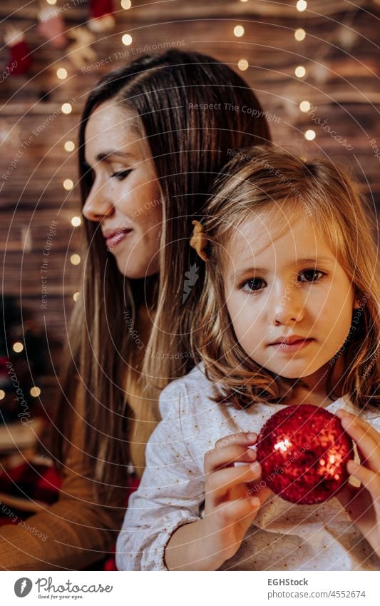 Hispanic Mutter hält ihre Tochter mit roten Ball und Lichter. . Wohnzimmer dekoriert durch Weihnachtsbaum und Geschenk Geschenk-Box, das Licht geben gemütliche Atmosphäre. New Year und Mama und Tochter Konzept