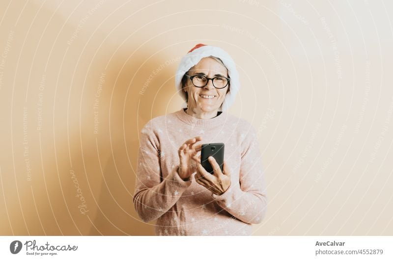 Happy Joyful Senior alten weißen Haaren Frau trägt Weihnachtsmann Hut lächelnd, während mit Smartphone. steht gegen eine Farbe Hintergrund. Wearing Weihnachten Kleidung. Kopierfläche. Werbefläche