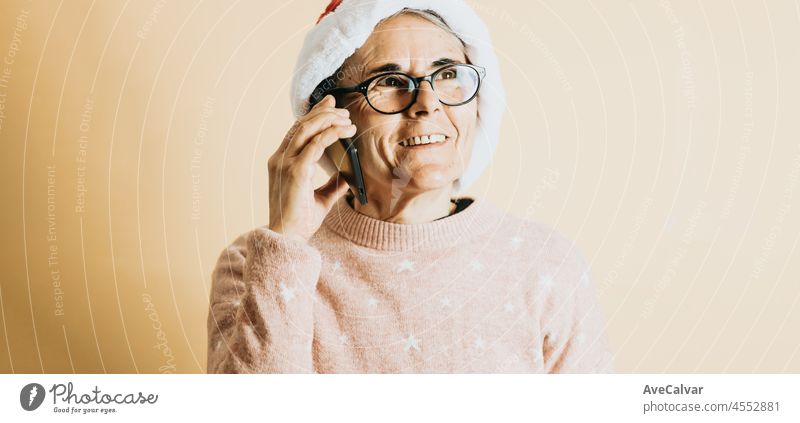 Happy Joyful Senior alten weißen Haaren Frau trägt Weihnachtsmann Hut lächelnd, während mit Smartphone. steht gegen eine Farbe Hintergrund. Wearing Weihnachten Kleidung. Kopierfläche. Werbefläche