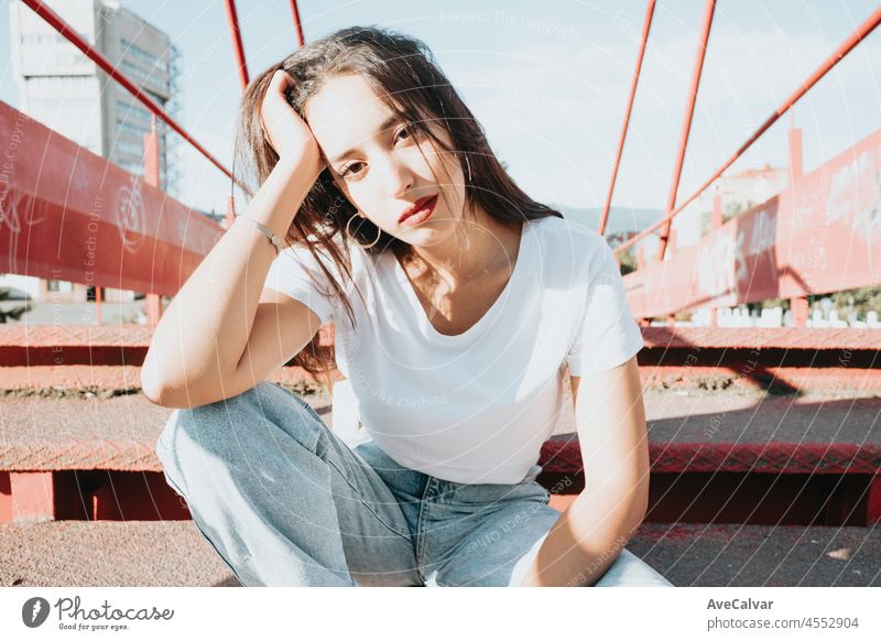 Urban Schuss eines modellierenden jungen afrikanischen Frau mit langen Haaren sitzen auf einem roten Boden schauen ernst in die Kamera. Stadt urbanes Konzept. Glücklicher Tag in der Stadt, weißes Hemd blaue Jeans. Modernes Outfit Styling.