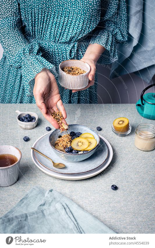 Frau gibt Müsli in eine Schüssel mit Obst hinzufügen Blaubeeren Kiwi Lebensmittel Frühstück Ernährung Portion Morgen Tee selbstgemacht Bestandteil