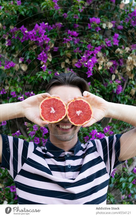 Fröhlicher Mann macht Augen mit Grapefruit und lächelt im blühenden Garten Lächeln Augen abdecken Freude Spaß haben Blütezeit positiv heiter Zitrusfrüchte