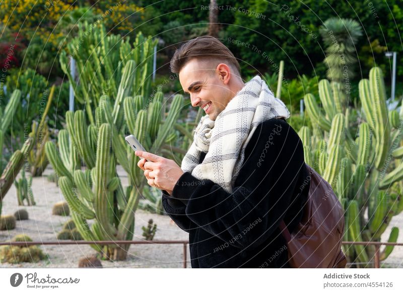 Positiver trendiger Typ, der in einem exotischen Garten auf seinem Smartphone surft Mann benutzend Lächeln positiv Browsen trendy tropisch Park charismatisch