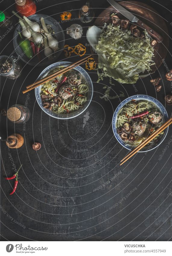 Essen Hintergrund mit traditionellen asiatischen Suppe in Schalen mit Stäbchen, Kohl, Knoblauch, Chili und Shiitake auf dunklen Betontisch. Würzige Ramen mit gesundem Gemüse zu Hause. Draufsicht mit Kopierbereich.
