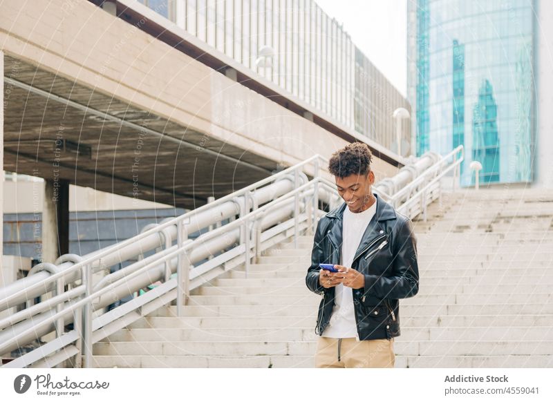 Schwarzer lächelnder Mann mit Smartphone auf Brückentreppe Funktelefon benutzend Texten Treppenhaus heiter Lächeln Schritt Laufmasche positiv Textnachricht