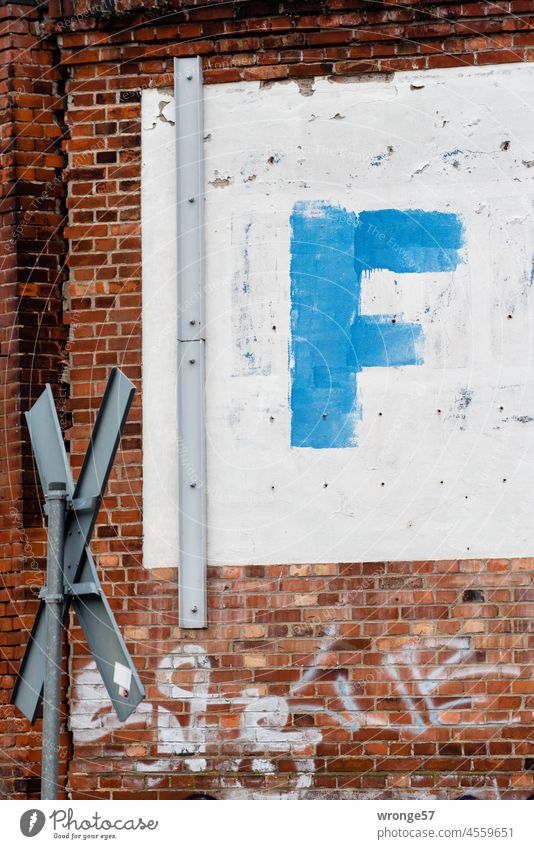 Ein großes blaues F auf weißem Hintergrund auf einer Backsteinmauer links von einem Andreaskreuz flankiert Buchstaben Großbuchstaben weißer Hintergrund Mauer