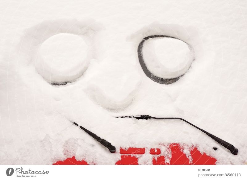 Auf der Frotscheibe eines eingeschneiten roten Autos sind zwei große Augen und eine Nase in den Schnee gemalt / Winter / Humor Neuschnee Schneemassen