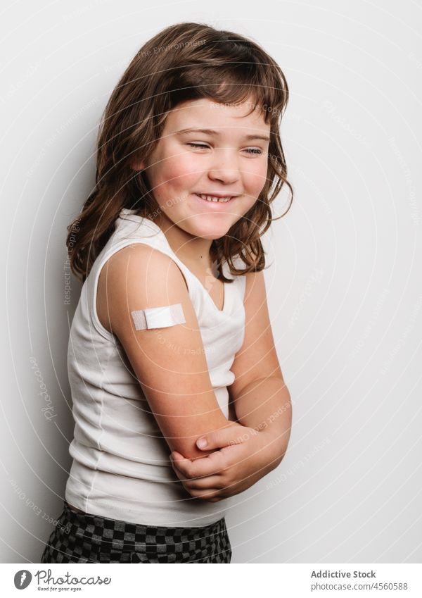 Glückliches geimpftes Mädchen mit Pflaster am Arm Schulmädchen bandagieren Band Impfstoff behüten heiter Virus Impfung Stoß Lächeln Schuss Sicherheit