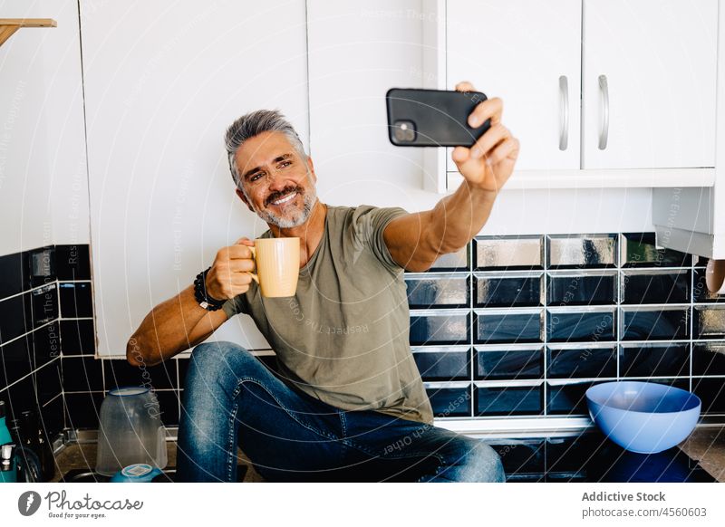 Bärtiger älterer Mann macht Selfie in der Küche Funktelefon zu Hause Smartphone Freizeit Lifestyle Abfertigungsschalter trinken Tasse Tee gutaussehend Kälte