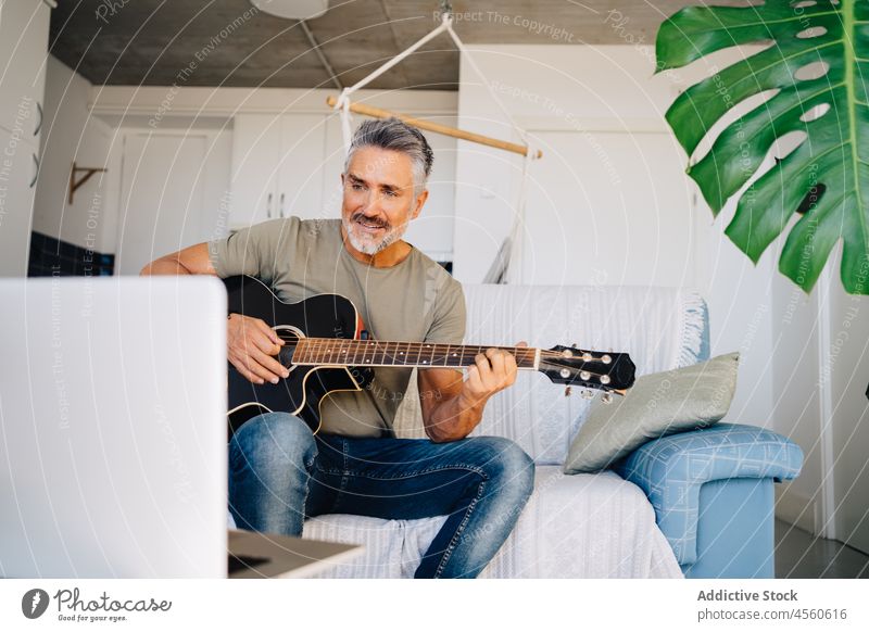 Älterer Gitarrist, der auf dem Sofa neben einem Laptop Musik macht Mann Gitarrenspieler Musiker Loft Instrument Innenbereich akustisch spielen online ausführen