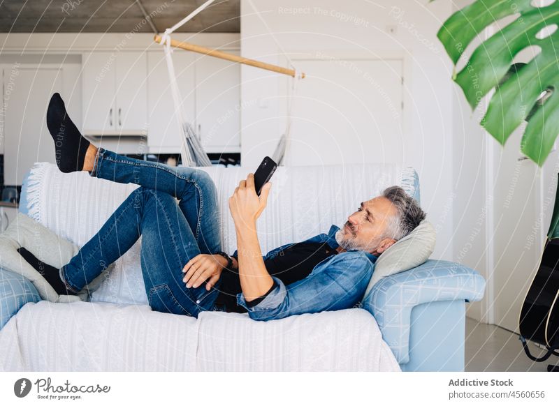 Älterer Mann, der auf der Couch liegend sein Handy durchsucht Smartphone Mobile Sofa Browsen benutzend Stil Loft Kälte ruhen Liege Zeitvertreib Funktelefon