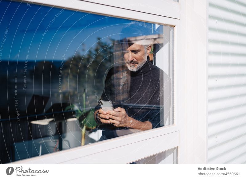 Älterer Mann, der ein Mobiltelefon hinter einem Glasfenster benutzt Fenster nachdenken Smartphone Funktelefon benutzend Browsen Zeuge neugierig achtsam Surfen