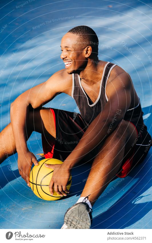 Lächelnder schwarzer Sportler auf dem Boden sitzend mit Ball im Stadion Mann Basketball Gericht Spieler Sportpark Sportbekleidung Athlet Aktivität Training