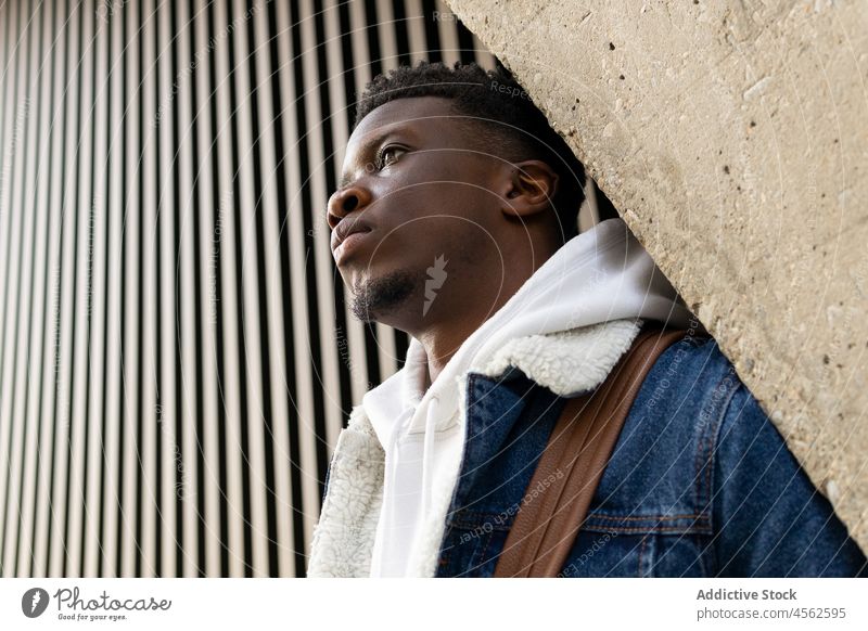 Stilvoller schwarzer Mann auf der Straße stehend und an die Wand gelehnt trendy Jeansstoff besinnlich Gebäude ernst Outfit nachdenklich Vorschein männlich