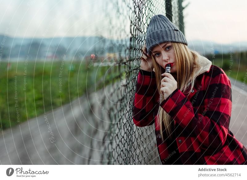 Junge Frau mit Wollmütze lehnt an einem Zaun und raucht Porträt jung Rauchen vaping alternativ in die Kamera schauen attraktiv Verdampfer schön blond Schlag