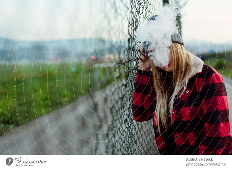 Junge Frau mit Wollmütze lehnt an einem Zaun und raucht jung Erwachsener vaping Rauchen alternativ gesichtslos attraktiv schön anonym blond Schlag Kaukasier