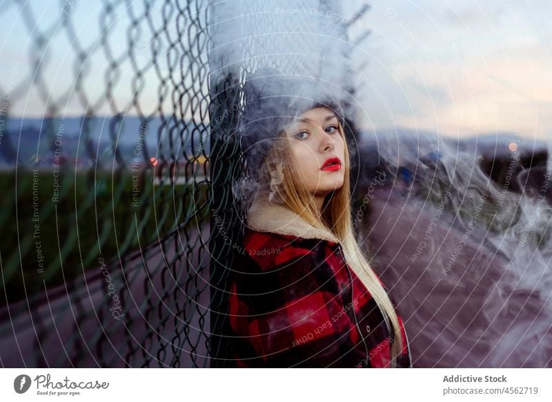 Junge Frau mit Wollmütze lehnt an einem Zaun und raucht jung Rauchen Porträt in die Kamera schauen Erwachsener vaping alternativ attraktiv schön blond Schlag