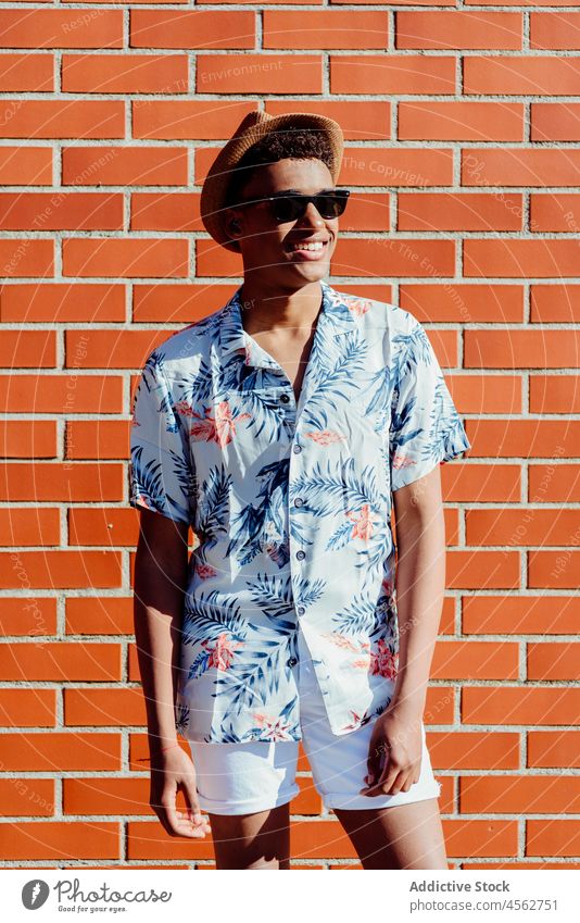 Trendiger junger schwarzer Mann posiert auf der Straße ethnisch Teenager Sonnenbrille Afrikanisch Hut Hemd geblümt Amerikaner Baustein Wand Porträt Stehen