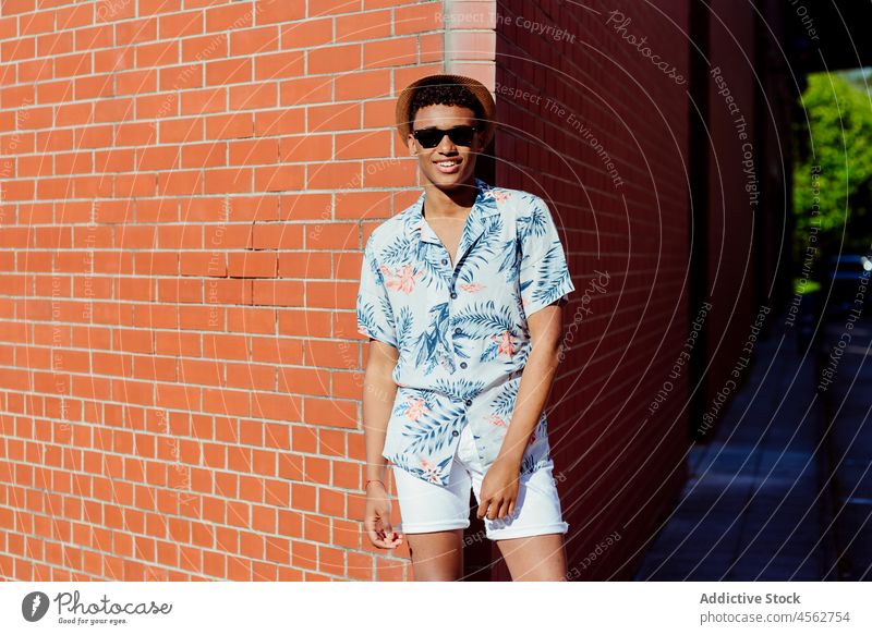 Trendiger junger schwarzer Mann posiert auf der Straße Teenager Sonnenbrille Hut Hemd geblümt Baustein Wand Porträt Stehen Großstadt urban posierend Aussehen
