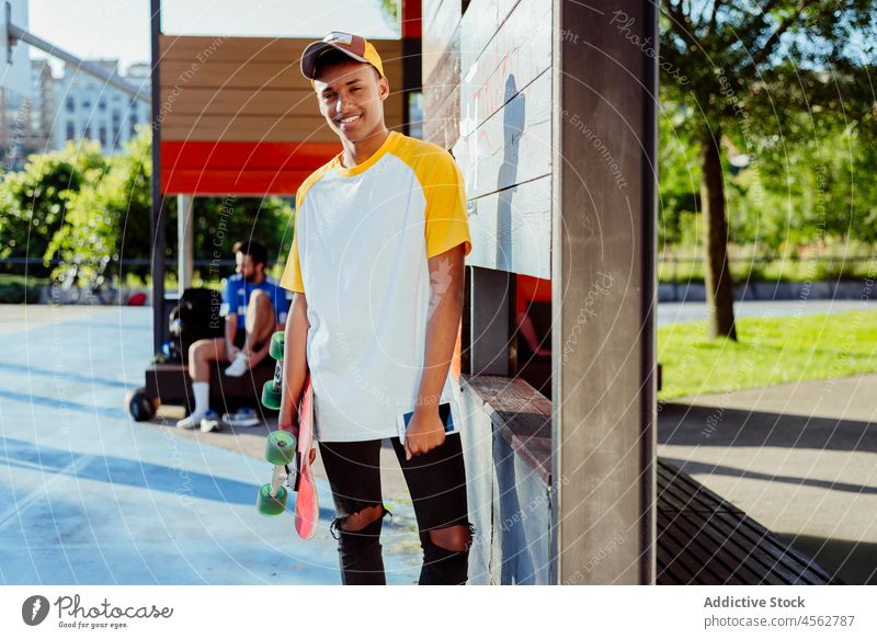 Junger schwarzer Mann mit T-Shirt und Mütze posiert im Park jung männlich Glück Lächeln Verschlussdeckel Telefon Straße Porträt urban Großstadt Skateboard