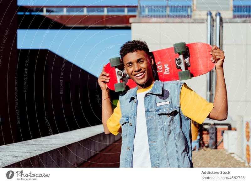Hübscher junger schwarzer Mann mit einem Skateboard auf der Straße männlich Junge Glück Lächeln Porträt urban Großstadt Baustein Wand Dachterrasse Weste