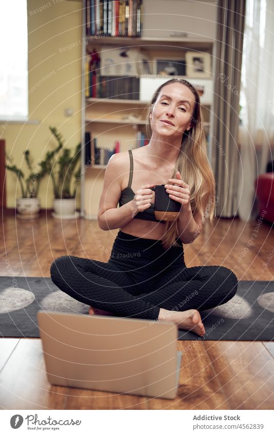Positive Frau mit heißem Getränk in der Nähe des Laptops während einer Yoga-Sitzung Training Gesunder Lebensstil Wellness Tutorial Lektion online Heißgetränk
