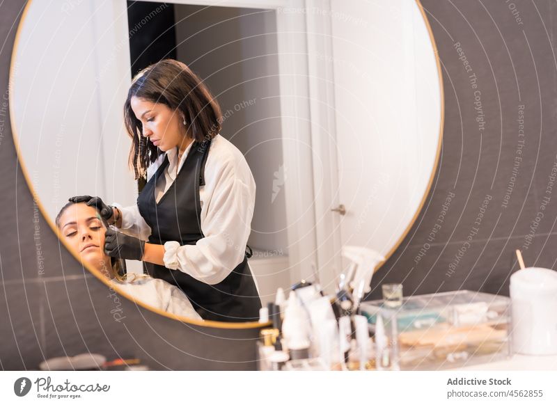 Kosmetikerin bürstet die Augenbrauen einer Kundin in einem Schönheitsstudio Frauen Salon Spiegel Klient Bürste Pflege Leckerbissen Reflexion & Spiegelung
