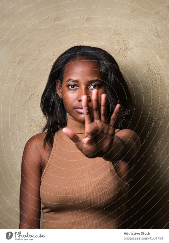 Ernste schwarze Frau steht mit erhobenem Arm vor einer beigen Wand ernst stoppen gestikulieren Zeichen Arm angehoben Konzentration begrenzen Aufmerksamkeit