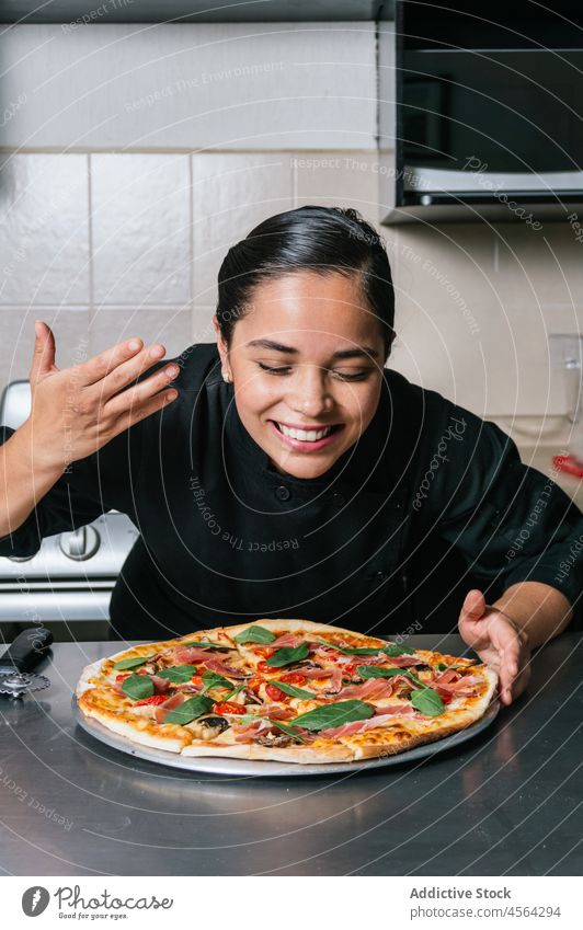 Lächelnde ethnische Köchin in Uniform genießt Pizzaduft Frau Mahlzeit Küchenchef Koch riechen Schniefen Lebensmittel geschmackvoll kulinarisch Speise Restaurant