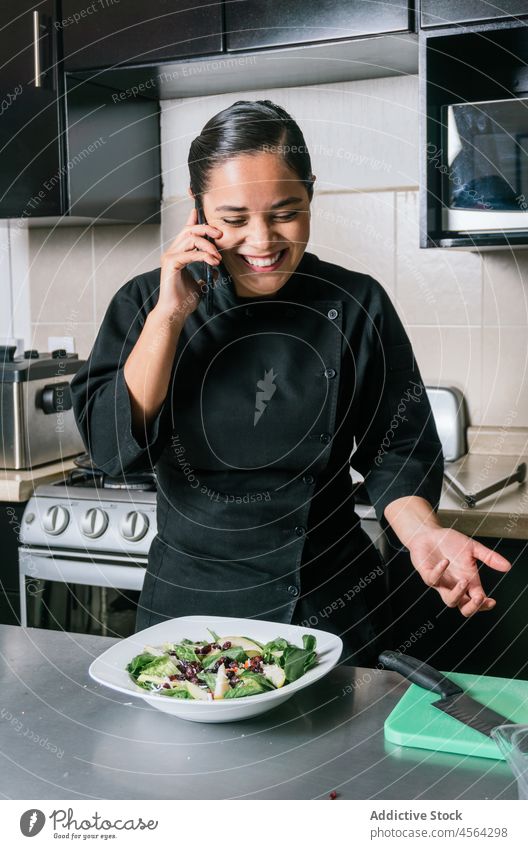 Lächelnder ethnischer Koch, der mit einem Smartphone spricht und einen Salat auf dem Tisch hat Frau Restaurant Küchenchef Speise Uniform kulinarisch reden