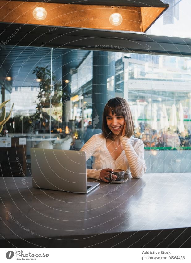 Fröhliche Frau mit einer Tasse Kaffee und Blick auf einen Laptop Videoanruf Café benutzend Browsen Gerät Anschluss abgelegen Gespräch freiberuflich Telefon