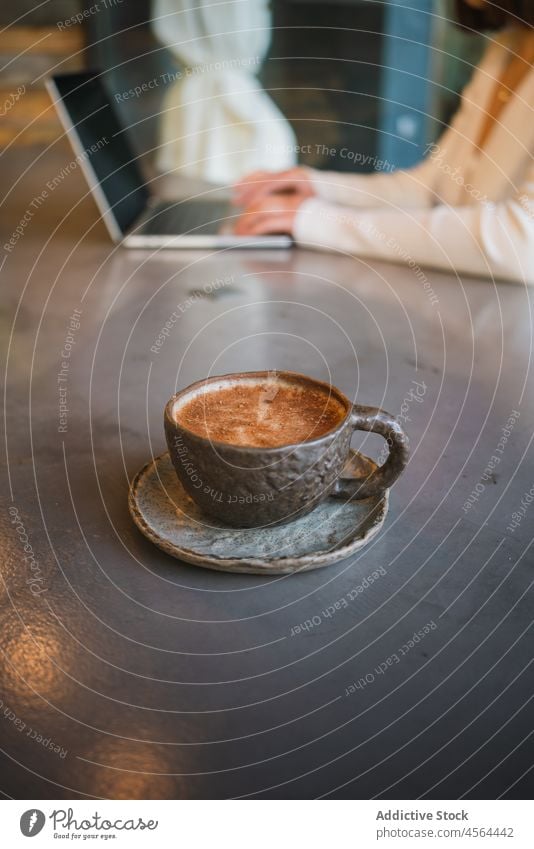 Eine Tasse Cappuccino steht auf dem Tisch neben einer Frau mit Laptop lecker Kaffee Apparatur trinken Tippen geschmackvoll benutzend freiberuflich Browsen Café