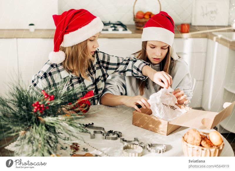 Zwei süße Mädchen mit Weihnachtsmannmützen sitzen am Küchentisch und verpacken süße Geschenke für Freunde und Verwandte. Ein Geschenk mit den eigenen Händen, eine süße Leckerei, festliche Vorbereitung